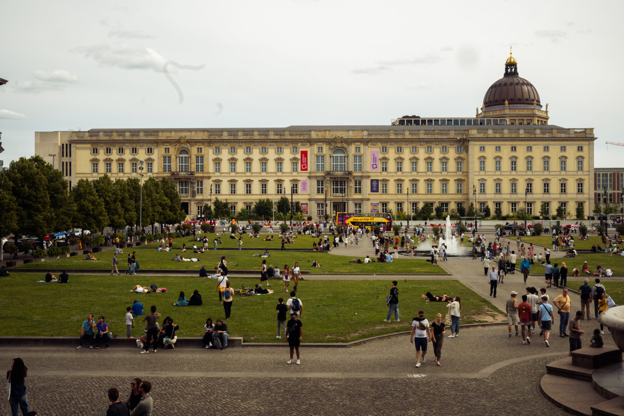 deSta_HF_Schloss - Dekoloniale Stadtführung Besucher entspannen auf dem Rasen vor dem Humboldt Forum, einem historischen Schloss mit Kuppel im Hintergrund.