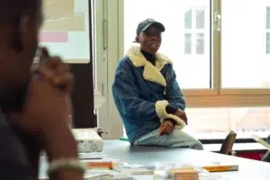 desta-workshop - Decolonial city tour Young person in blue jacket sitting at a table in a workshop, looking at the camera, presentation in the background.