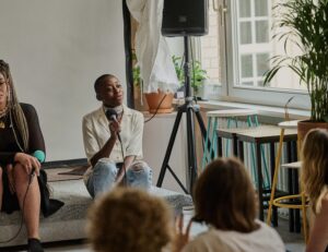 Speaker_Job_Antiracism_Work - Decolonial city tour Speaker with microphone sitting on a couch during a discussion about anti-racism work in a bright room.