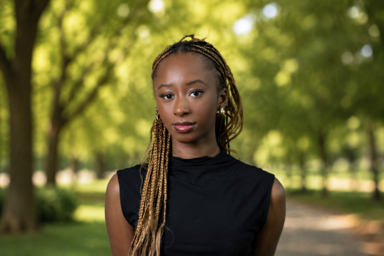 A young woman with braided hair is standing in a green park, surrounded by trees.